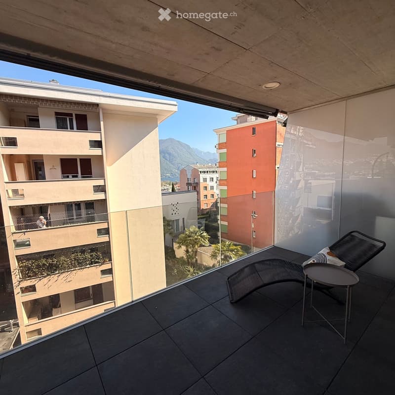 Modern balcony with dark tiled floor, glass railing, and a view of the city and mountains. Includes a lounge chair and a small table.