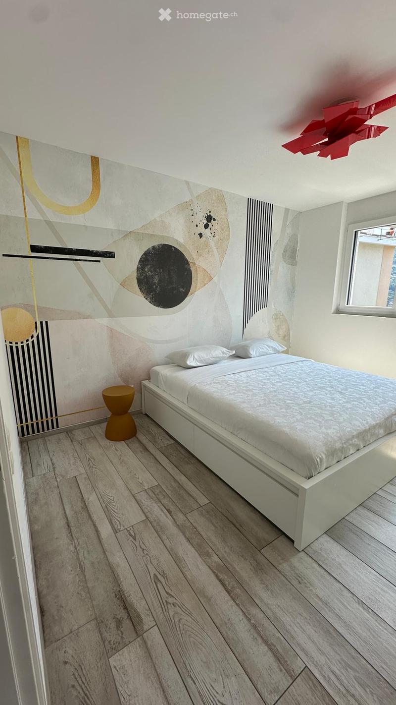 Modern bedroom with a white bed, two pillows, a small brown stool, and a large abstract wall mural. The room features light wooden flooring and a red geometric light fixture on the ceiling.