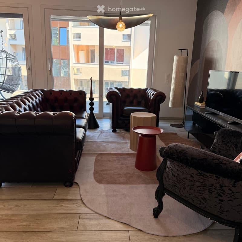 Modern living room with a large window and balcony access, featuring a dark leather sofa, a matching armchair, a glass coffee table, a red side table, a television on a stand, and a hanging wicker chair near the window.