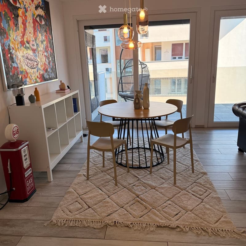 Modern living room with wooden floor, round dining table, six chairs, shelf with decorative items, gas station prop, and large painting on wall. Balcony access visible.