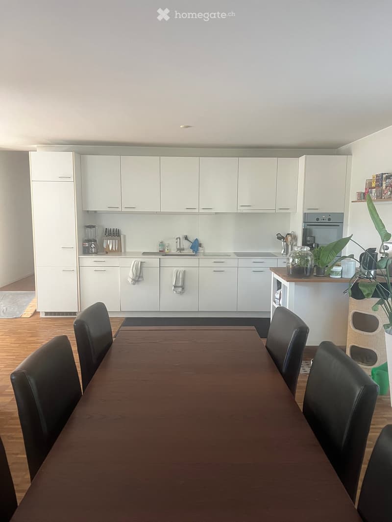 Modern kitchen with white cabinets, a wooden dining table with black chairs, and various appliances including a blender, knife block, and oven. A small plant is placed on the counter.