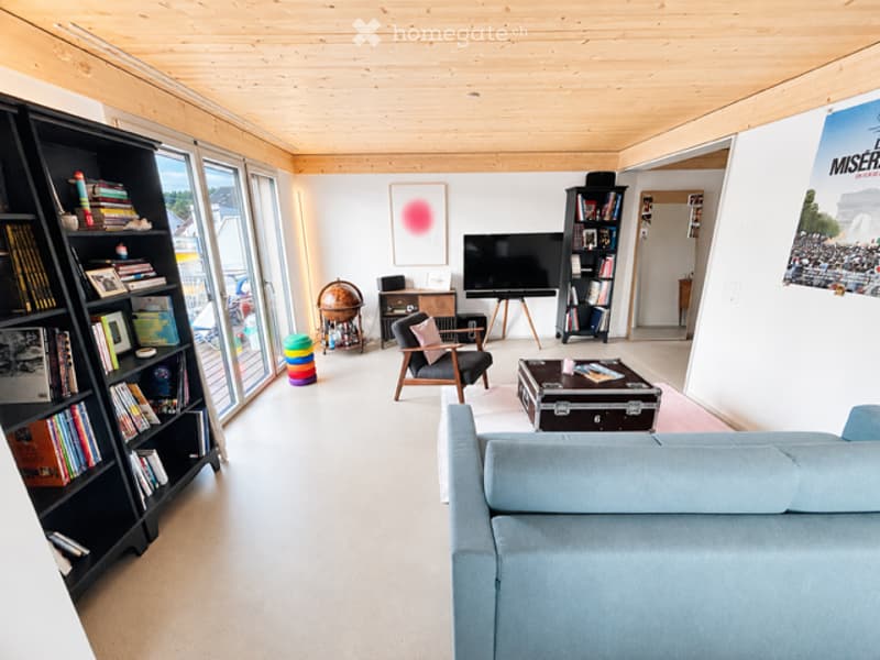 Bright living room with wooden ceiling, large windows, sofa, coffee table, bookshelf, chair, and artwork.