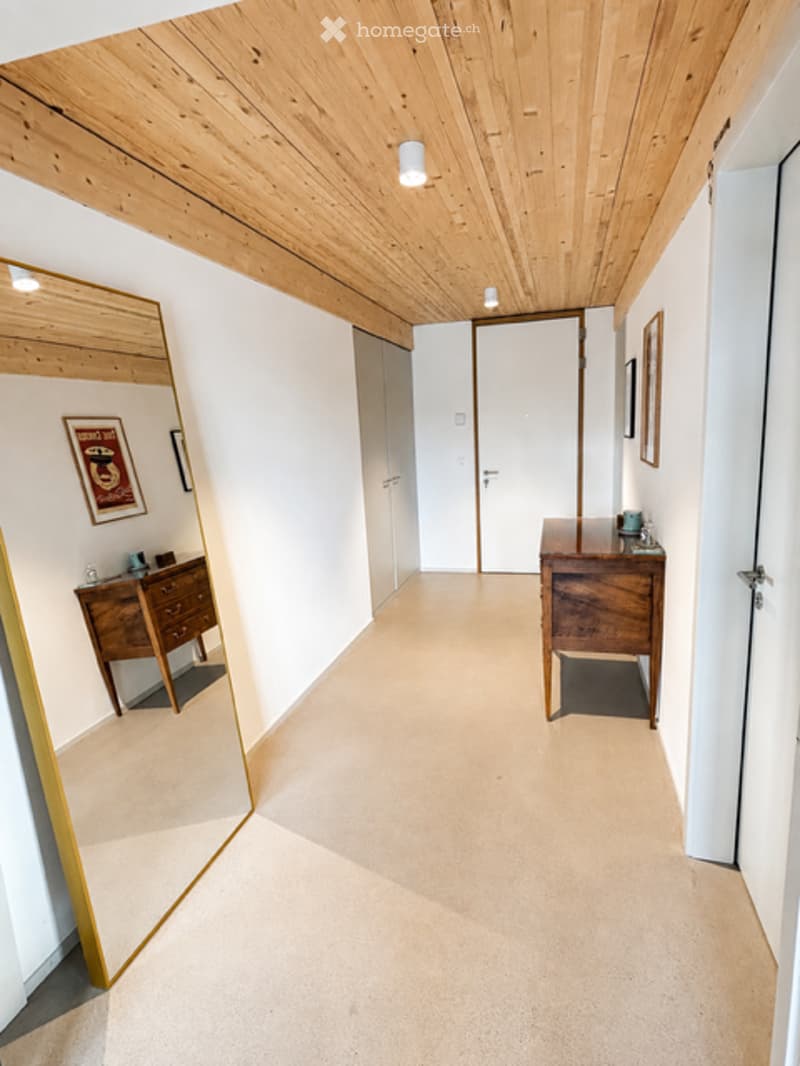 A modern hallway with light wooden ceiling, white walls, and a large mirror on the left side. There are wooden dressers with small items on top and framed artwork on the walls.