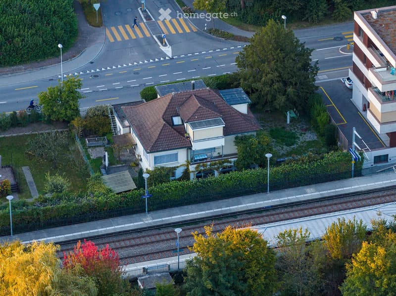 Aerial view of a residential area with a two-story house, a multi-story apartment building, a railway track, and surrounding greenery.