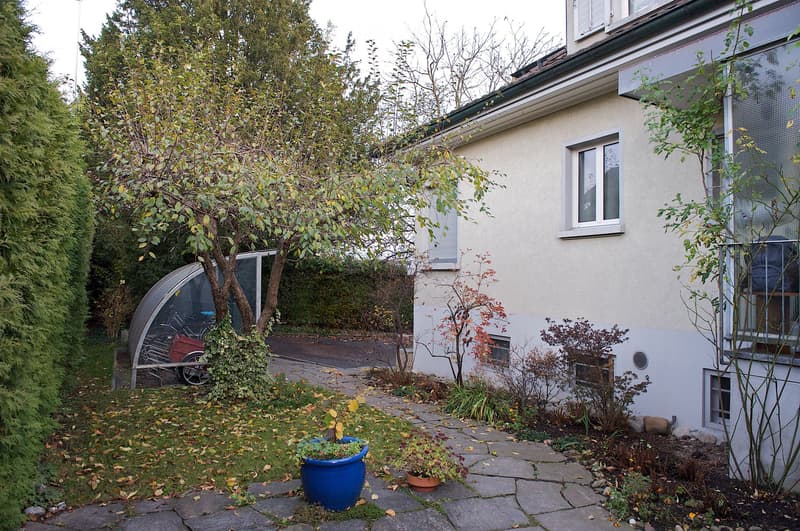A well-maintained garden area with a paved stone pathway, potted plants, and a modern house with large windows and a curved glass structure.
