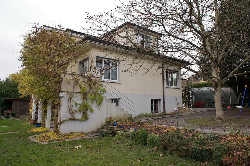 Two-story house with beige and gray exterior, multiple windows, and a staircase leading to an upper level. The surrounding area includes a garden with grass, trees, and a small play area with a swing and slide.