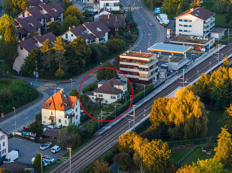 Aerial view of a residential area with multiple houses, a train station, a gas station, and surrounding greenery.