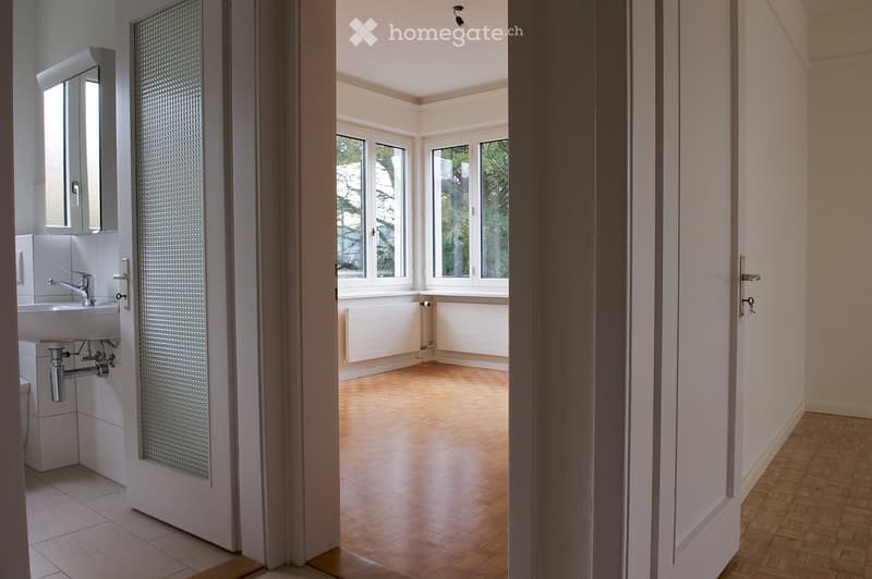 Empty room with wooden flooring, large windows, and a partially visible bathroom area with a sink and mirror.