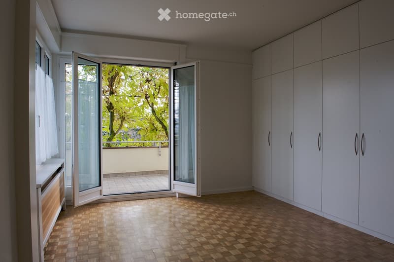 Empty room with wooden flooring, white built-in wardrobes, large sliding glass doors opening to a balcony, and white walls.