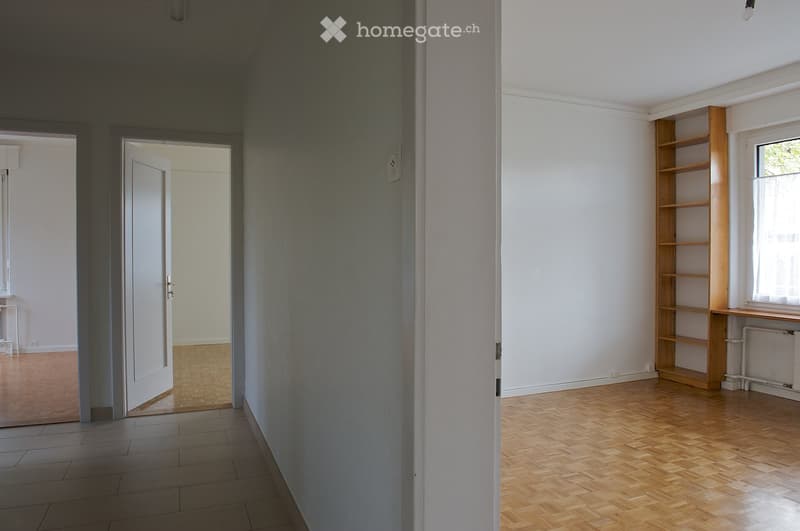 Empty room with wooden flooring, white walls, and a wooden shelf unit near a window. The room has multiple doors leading to other spaces.
