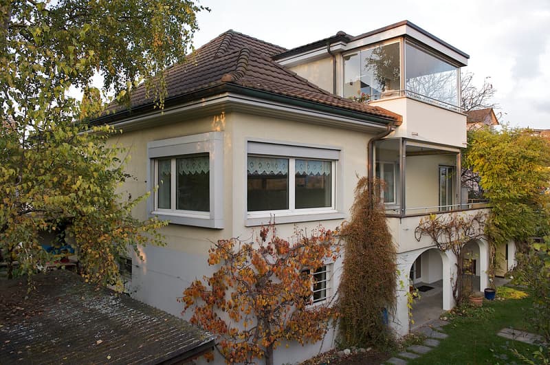 Two-story house with a tiled roof, large windows, and a balcony on the upper level. The house is surrounded by trees and has a small garden area with a stone pathway.