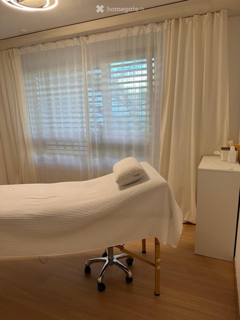 A well-lit room with a massage table covered in a textured white sheet, a small pillow on top, and a small cabinet with toiletries visible on the right side.