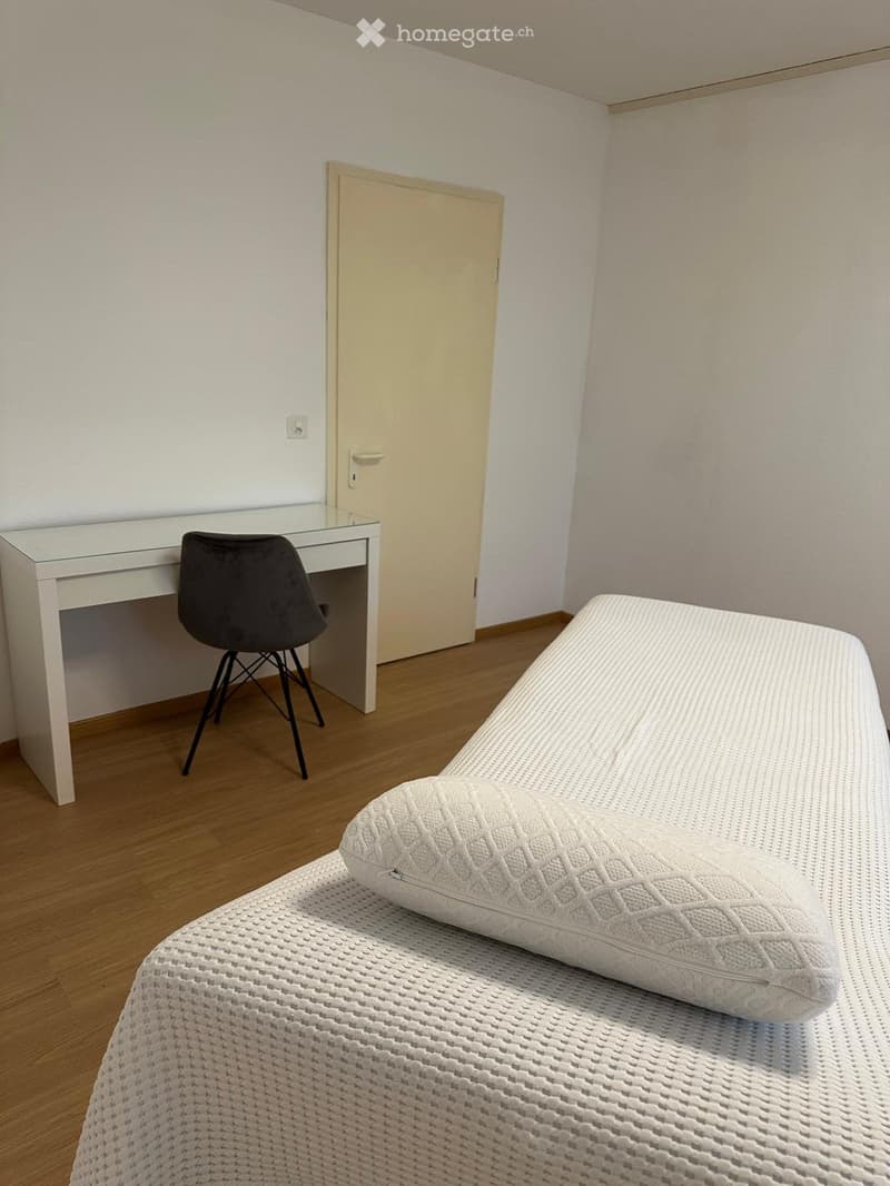 Modern bedroom with a white bed, a white desk, and a black chair. The room features light-colored walls and wooden flooring.