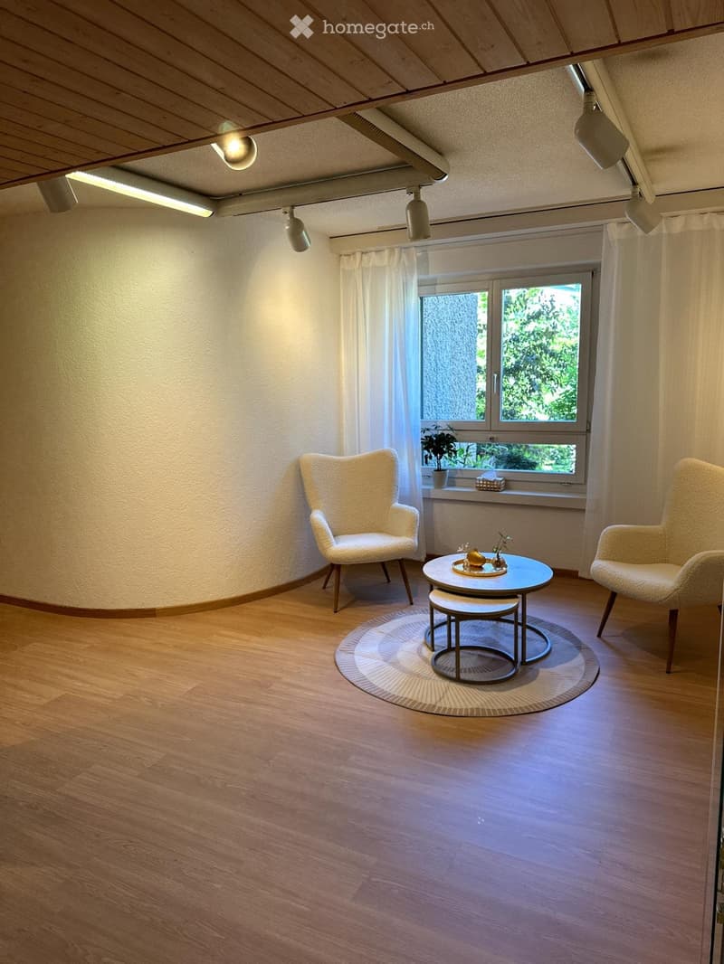 Bright living room with wooden flooring, two white chairs, a round coffee table with two stools, a round rug, and a small plant on the windowsill.