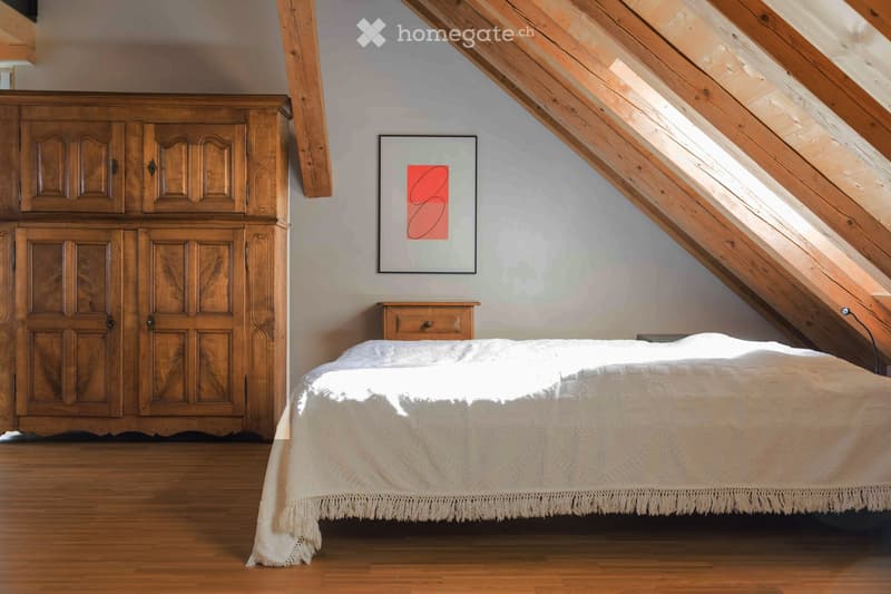 Cozy attic bedroom with wooden slanted ceilings, a large wooden wardrobe, a neatly made bed with a white duvet, a small nightstand, and a modern abstract artwork on the wall.