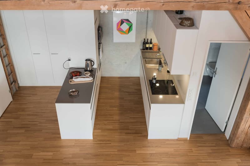 Modern kitchen with white cabinets, wooden flooring, and stainless steel appliances. Features an induction stove, sink, and a small bathroom visible in the background.