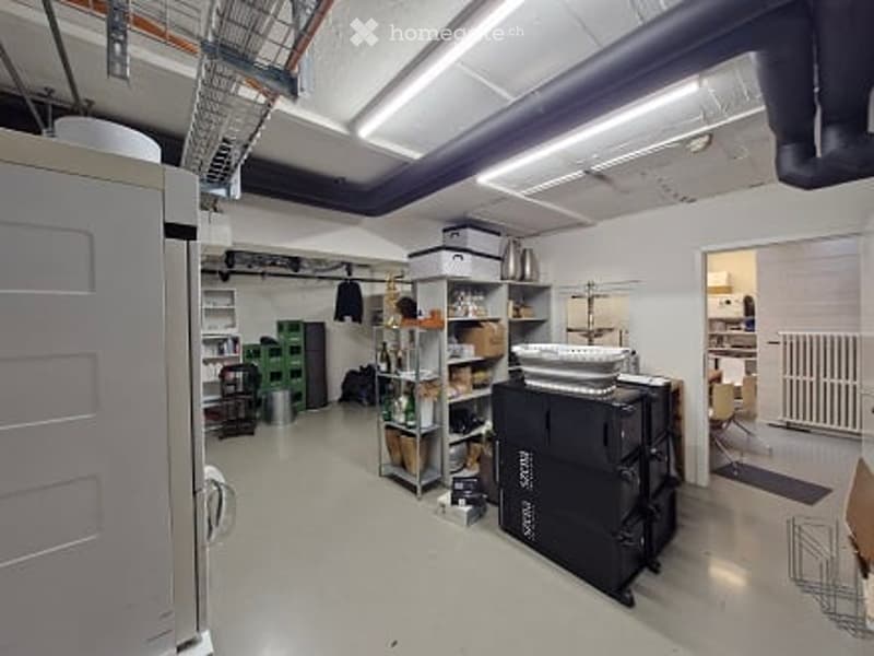 The image shows a spacious, well-lit room with industrial-style lighting and exposed ductwork. The room contains a refrigerator on the left, shelves with various items, storage boxes, a table with a bowl on it, and a chair. The floor is concrete, and the