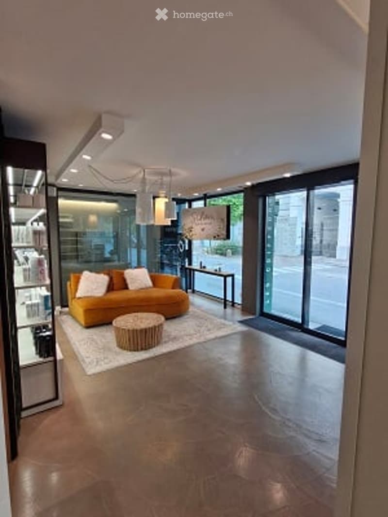 Modern living room with large glass windows, a yellow sofa, a round coffee table, and a floor lamp. The room features a polished concrete floor and a white rug.