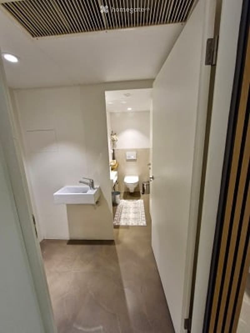 Modern bathroom with a white sink, chrome faucet, toilet, and shower area. The space features light-colored tiles and a sleek design.