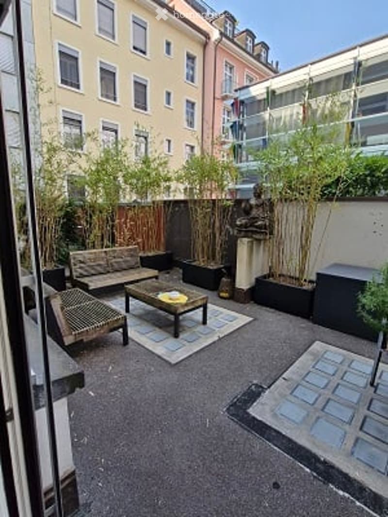 Courtyard with a seating area, including a bench, small table, and chairs, surrounded by potted plants and a decorative statue.