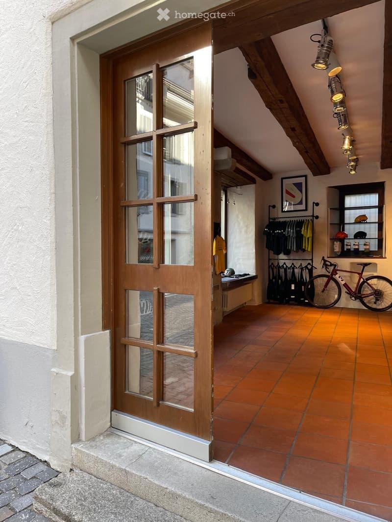 A small, well-lit room with exposed wooden beams, terracotta tiles, and a wooden door with glass panels. Contains a bicycle, a clothing rack, a shelf, and a picture frame.