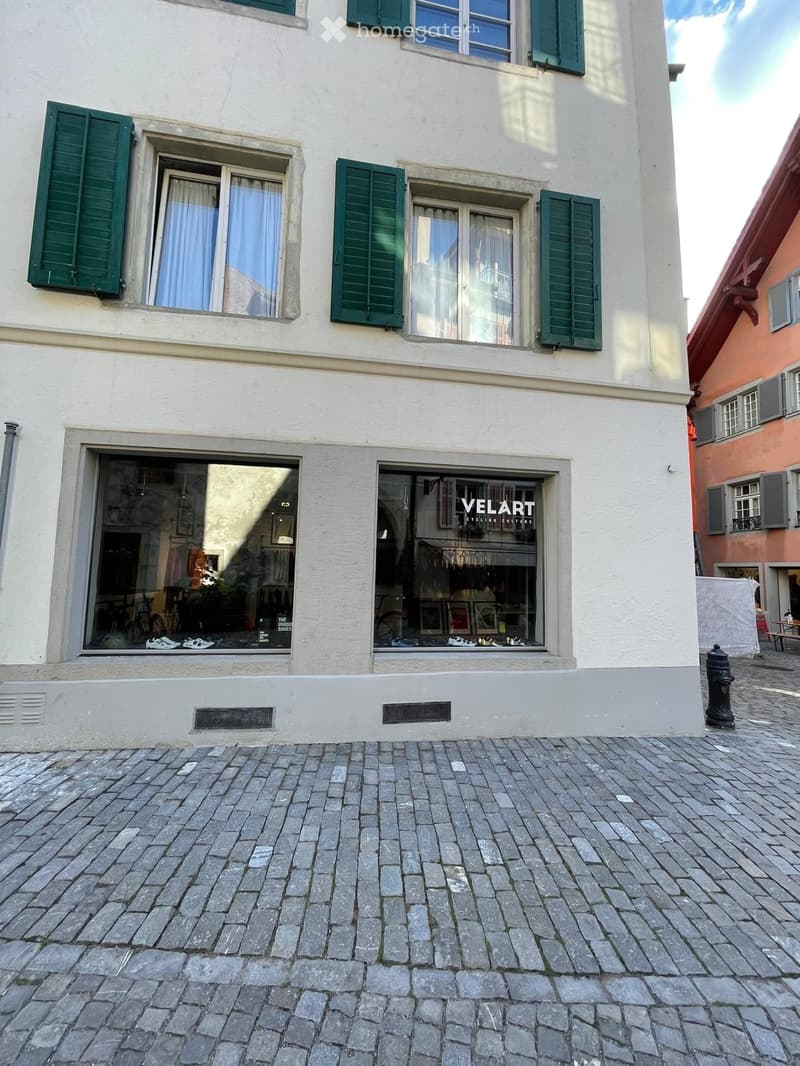 Two-story building with white facade, green shutters, and large windows. Ground floor features two storefronts with glass displays showing shoes and cycling gear.