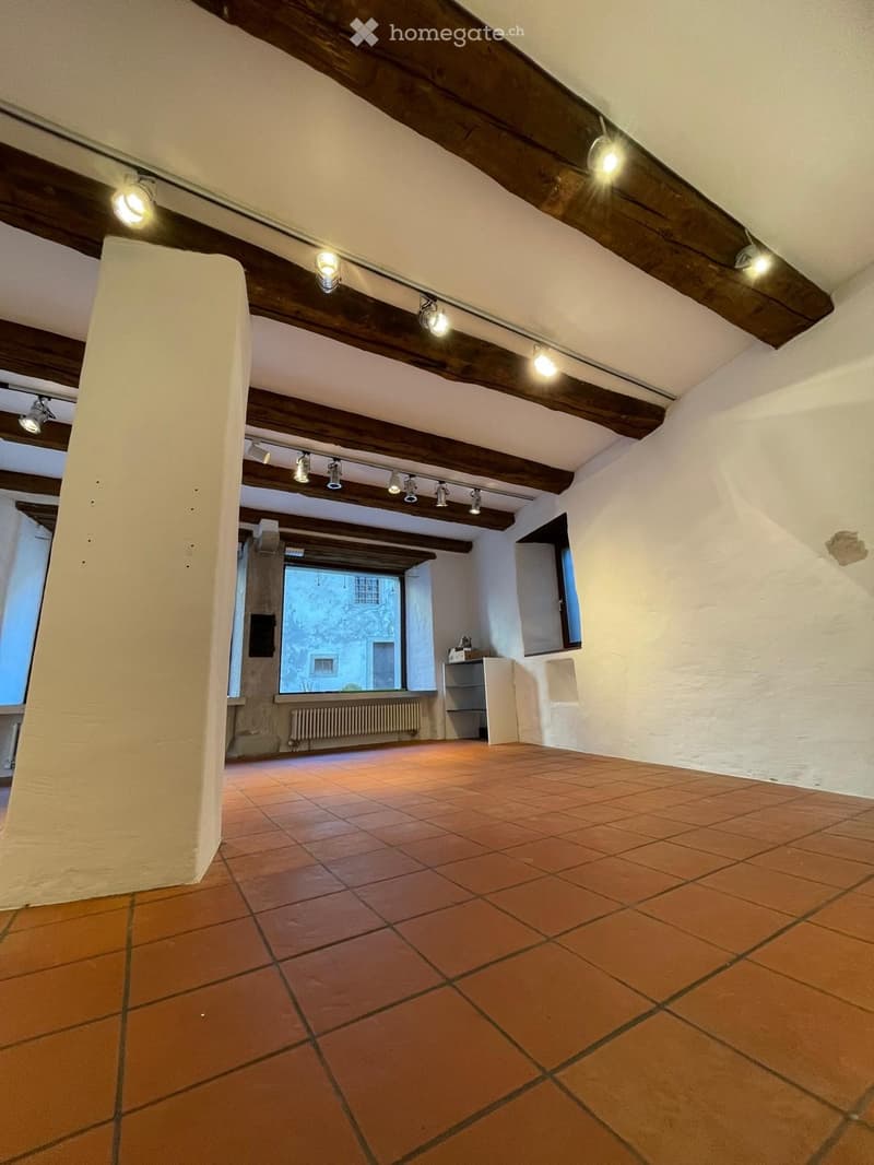Empty room with exposed wooden beams, terracotta tiled floor, white walls, and large windows offering a view of the outside.