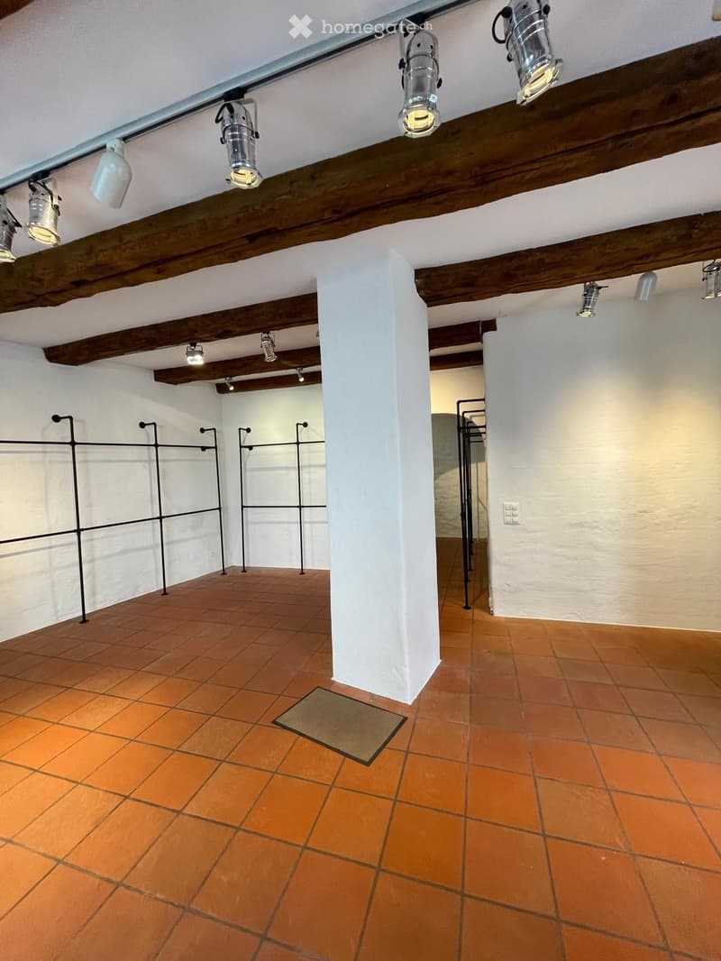 Empty room with exposed wooden beams, terracotta tiles, and white walls. Features a central pillar and several hanging spotlights.