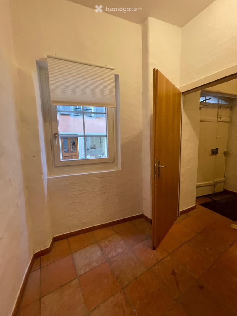A narrow hallway with tiled flooring, a window with blinds, and a wooden door leading to another room.