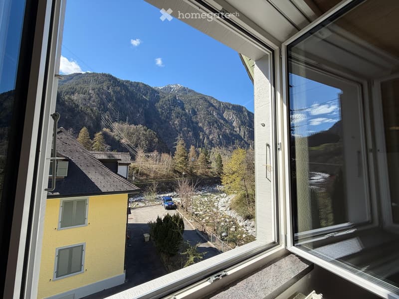 View from a balcony showing a scenic mountain landscape, a yellow house, and a parked car below.
