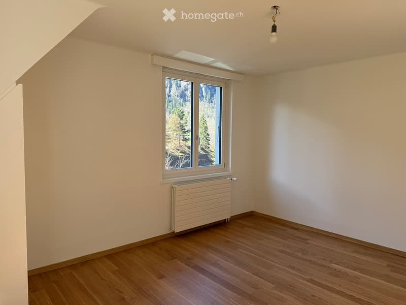 Empty room with wooden flooring, white walls, a window with a view of trees, a radiator, and a ceiling light fixture.