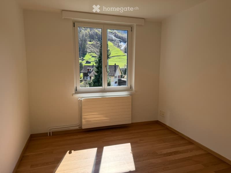 Empty room with wooden flooring, large window with scenic view, radiator, and light switch.