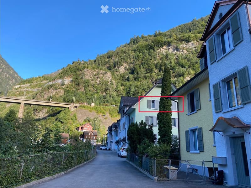 A row of residential buildings in a picturesque village with a mountainous backdrop, featuring a mix of architectural styles and a clear blue sky.