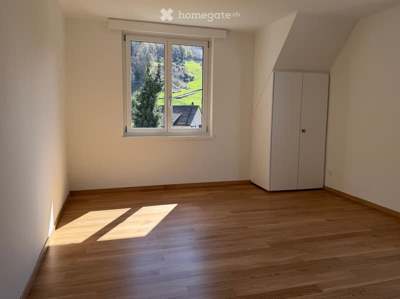 Empty room with wooden flooring, white walls, and a large window offering a view of greenery outside. A built-in closet is visible on the right side.