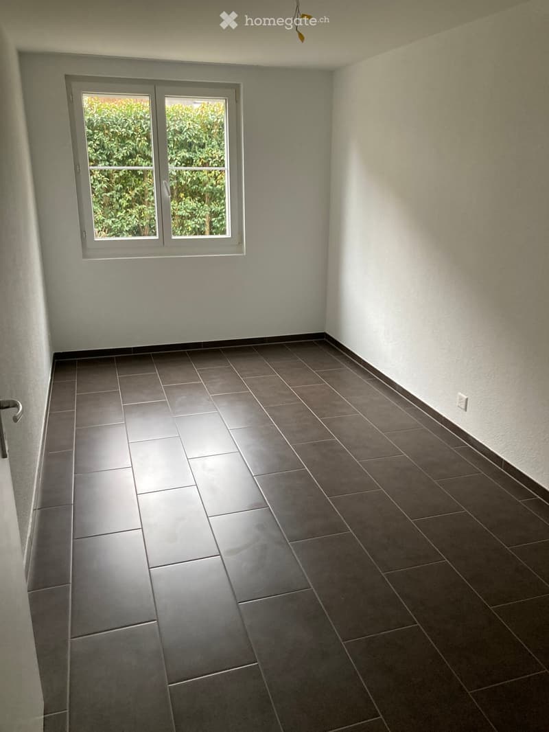 Empty room with tiled floor and white walls, featuring a large window with a view of greenery outside.
