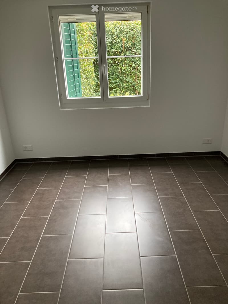 Empty room with grey tiled floor and light grey walls, featuring a large window with green shutters and a view of greenery outside.