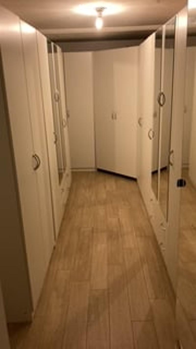 A long, narrow hallway with light wooden flooring and white walls. The corridor features multiple closed white doors on both sides, with a single ceiling light fixture providing illumination.