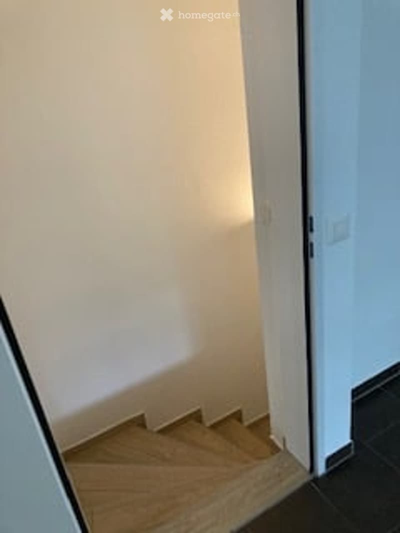 A narrow hallway with wooden stairs leading up, white walls, and a dark tiled floor.