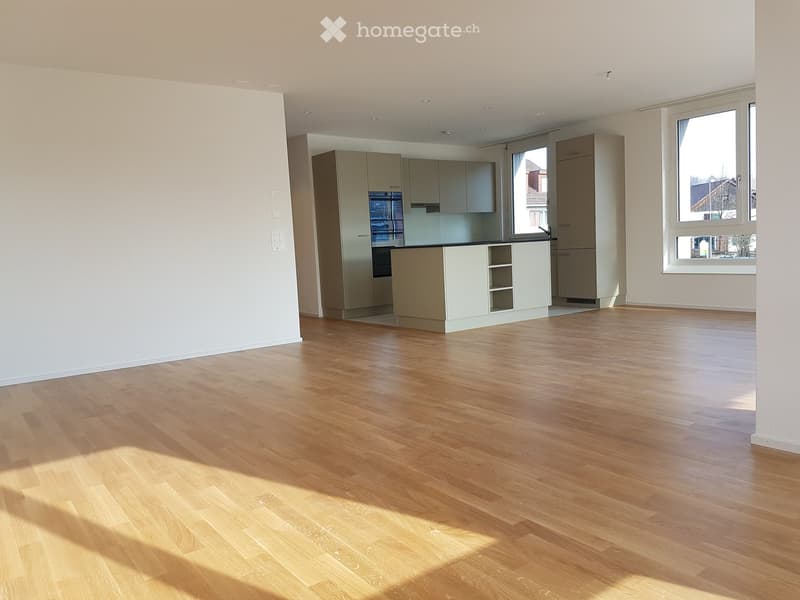 Empty room with light wooden flooring, white walls, and a small kitchen area with cabinets and a countertop visible in the background.
