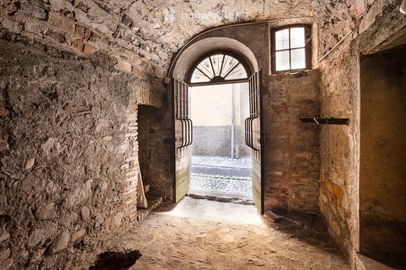 A narrow, stone-walled corridor with an arched metal gate leading to an outdoor area. The walls are made of rough stone and brick, and the floor is unpaved.