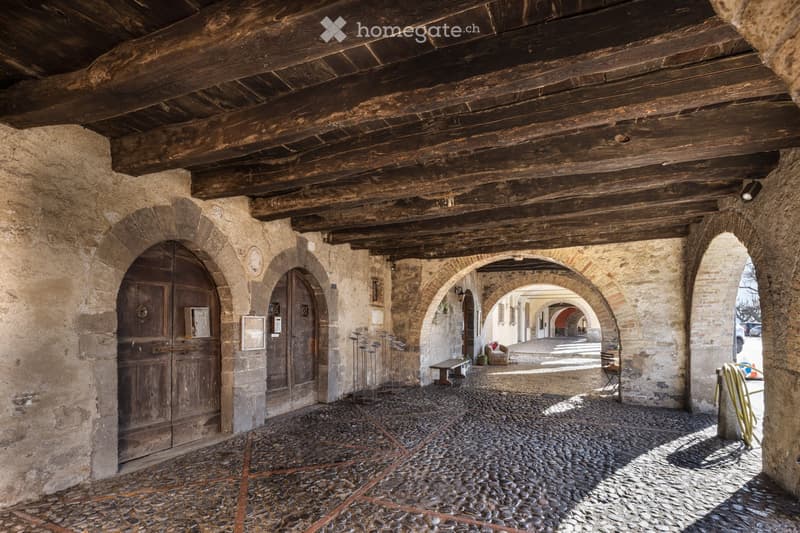 A covered stone corridor with wooden beams, cobblestone floor, arched doorways, and minimal furnishings including a small table and chair in the distance.