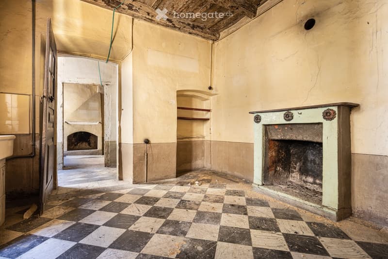 Empty room with exposed wooden beams, checkered tile floor, fireplace, built-in shelves, and a doorway leading to another room.