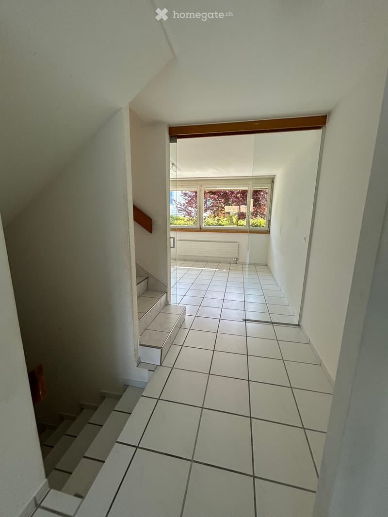 A narrow, tiled hallway with white walls, leading to a room with large windows and a radiator. The hallway has a staircase on the left side.
