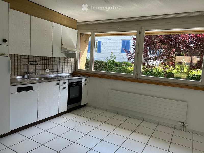 Modern kitchen with white cabinets, tiled backsplash, stainless steel sink, and appliances including a refrigerator, dishwasher, and oven. Large windows offer a view of the garden.