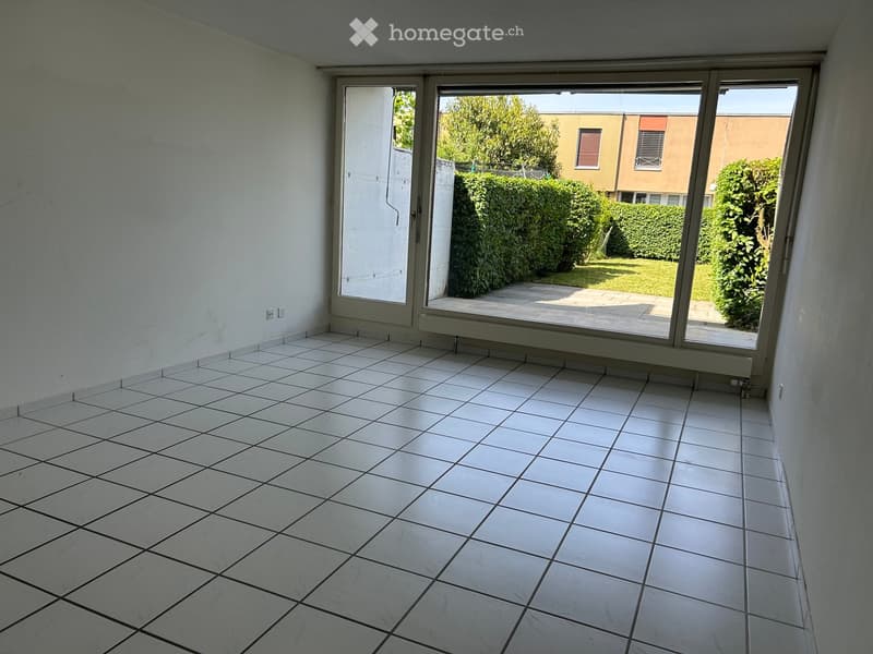 Empty room with white tiled floor and large sliding glass doors leading to a patio area with greenery visible outside.