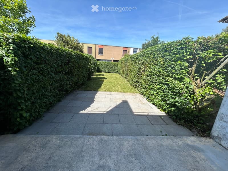 A paved garden pathway bordered by tall hedges and greenery, leading to a grassy area with a building visible in the background.