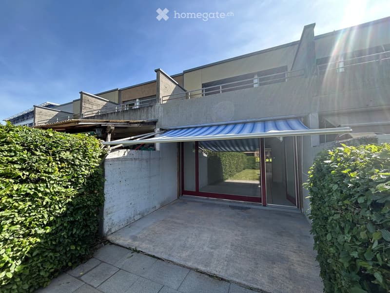 Modern multi-story building with a concrete facade, large glass windows, and a covered entrance with striped awning. The entrance leads to a spacious patio area surrounded by neatly trimmed hedges.