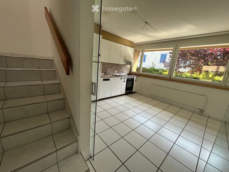Empty kitchen with white tiled floor, white cabinets, and a window overlooking a garden. No furniture or appliances are present.