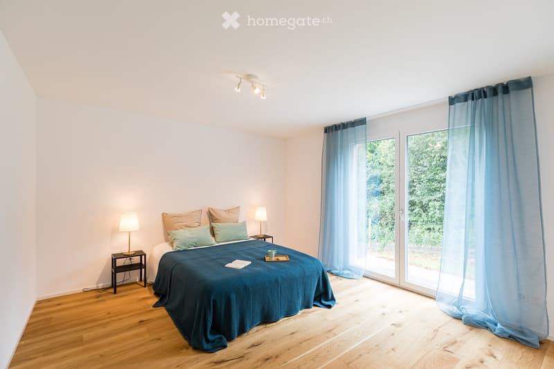 Bright bedroom with wooden flooring, large bed with blue bedding and multiple pillows, two nightstands with lamps, and large windows with blue curtains leading to a balcony.