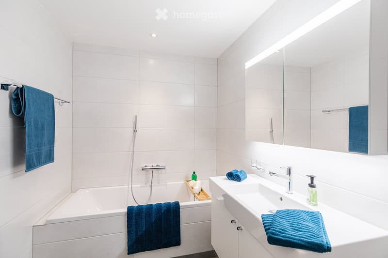 Modern bathroom with white tiles, a bathtub, a sink, a large mirror, and blue towels.
